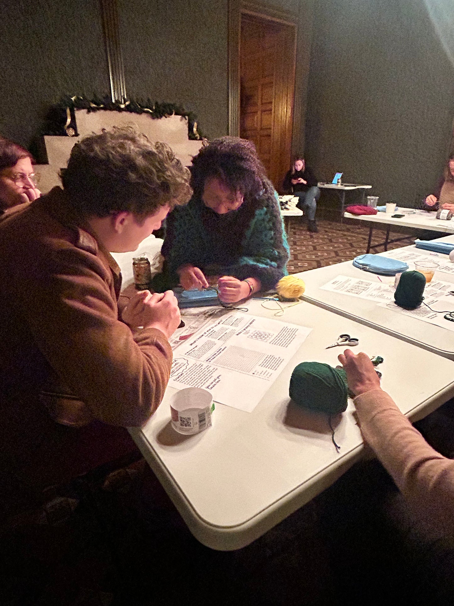 A person with kinky hair and caramel colored skin and a green and black crochet sweater holds yarn over a white table, while students get more detail on how to crochet. A guide is on the table.