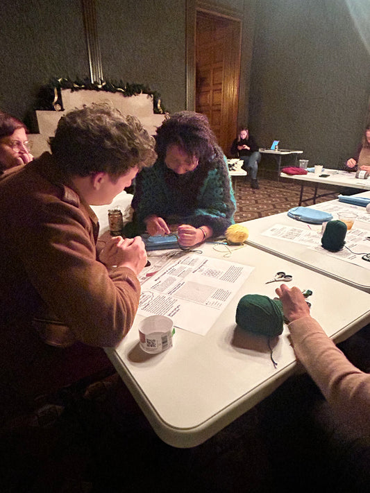 A person with kinky hair and caramel colored skin and a green and black crochet sweater holds yarn over a white table, while students get more detail on how to crochet. A guide is on the table.